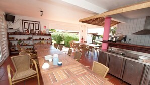 An outdoor dining area with a long wooden table, wicker chairs, and a kitchen area with stainless steel appliances. - Jéricho Gîtes & Spa (Capesterre-de-Marie-Galante)