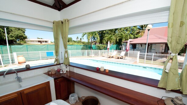 A bathroom with a large window overlooking a pool area, wooden cabinets, a sink, and a towel on the counter. - Jéricho Gîtes & Spa (Capesterre-de-Marie-Galante)