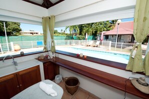 Une salle de bain dotée d’une grande fenêtre donnant sur une piscine, de meubles en bois, d’un lavabo et d’une serviette posée sur le comptoir.