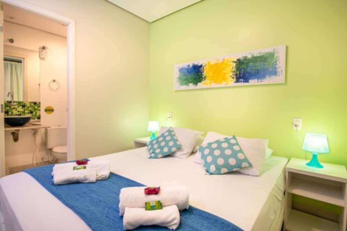 A hotel room with a bed, two nightstands, a wall painting, and a bathroom visible through an open door.