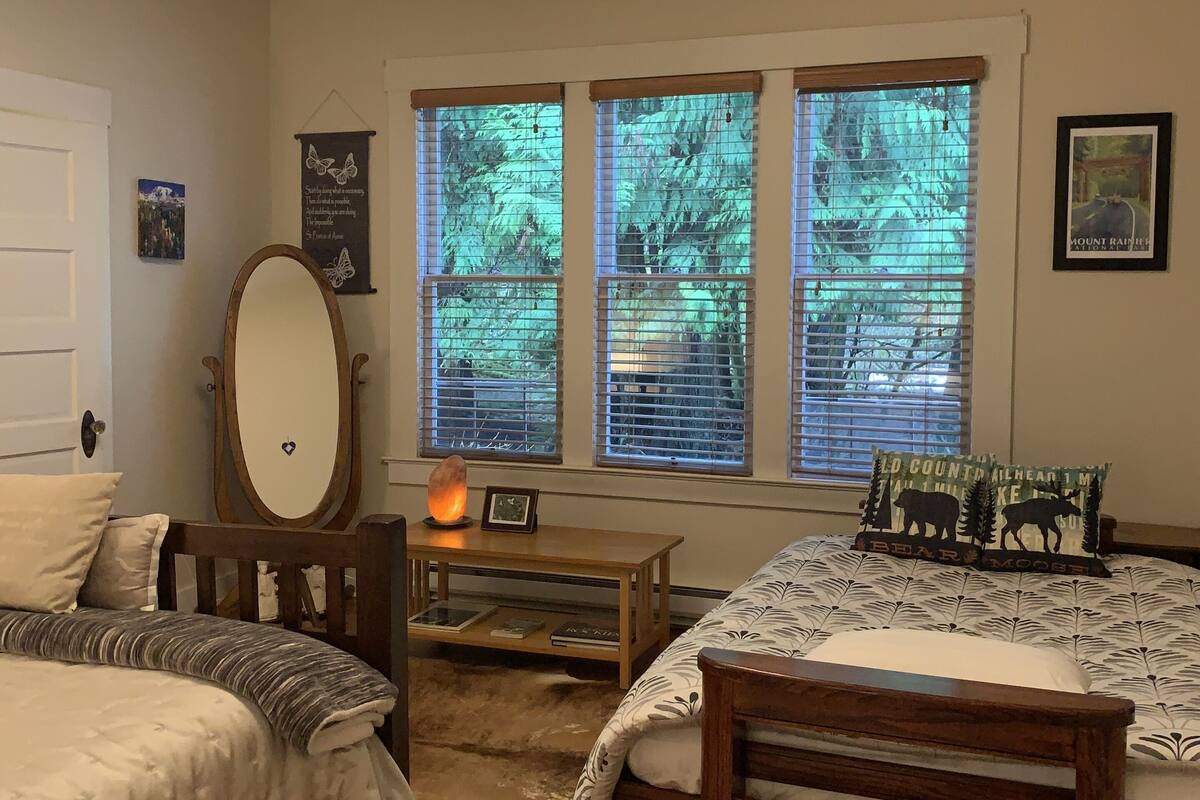 A bedroom with two beds, a wooden headboard, a mirror, a bedside table with a lamp, and a window with blinds.
