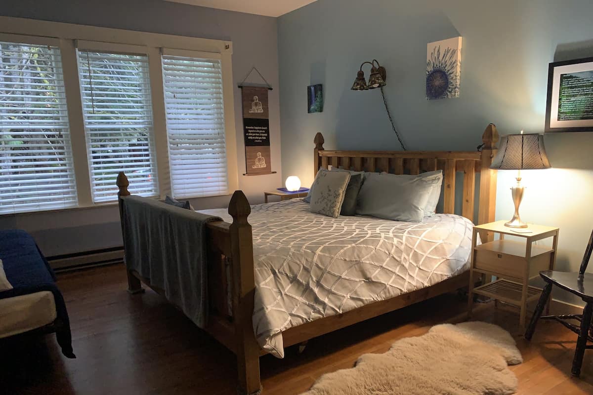 A bedroom with a wooden bed, a nightstand with a lamp, a chair, and a large window with blinds.