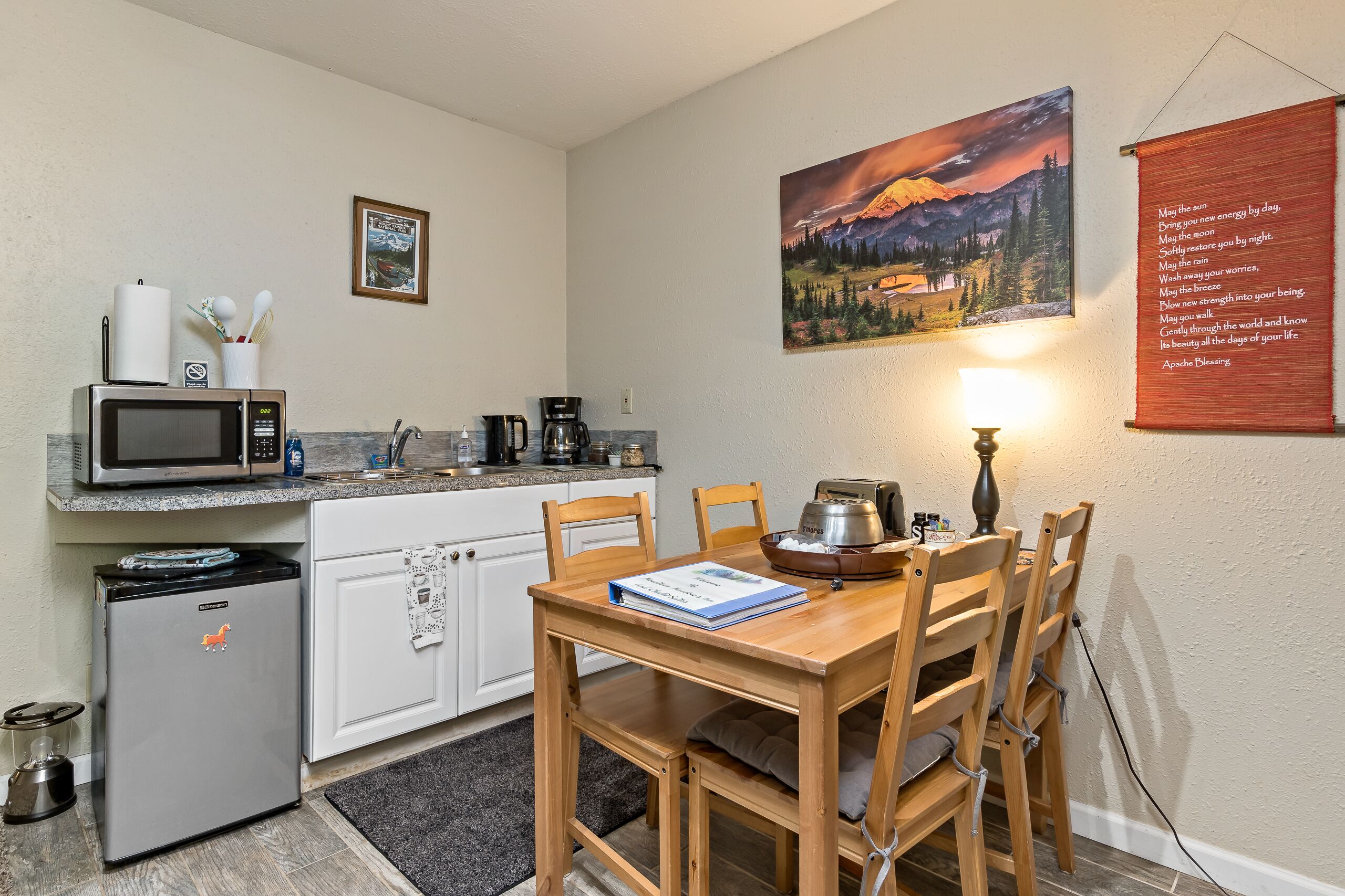 A compact kitchen with a microwave, coffee maker, and a small refrigerator. A dining table with chairs is positioned under a painting of a mountain landscape.