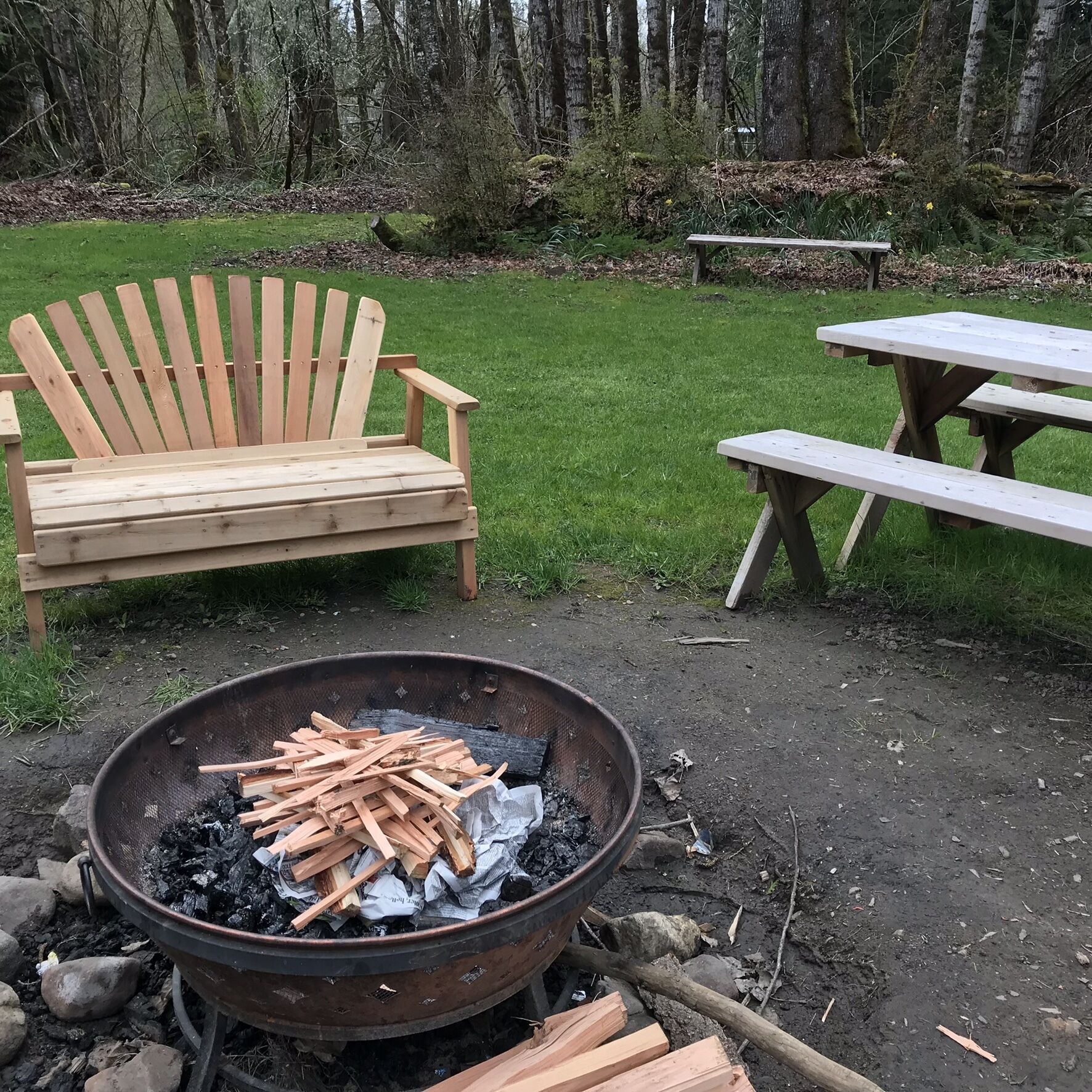A wooden Adirondack chair and a picnic table in a grassy outdoor area with a fire pit in the foreground.