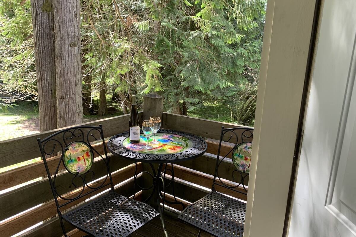 A round table with a colorful glass top, two metal chairs, and a bottle of wine on a wooden deck with a view of trees.