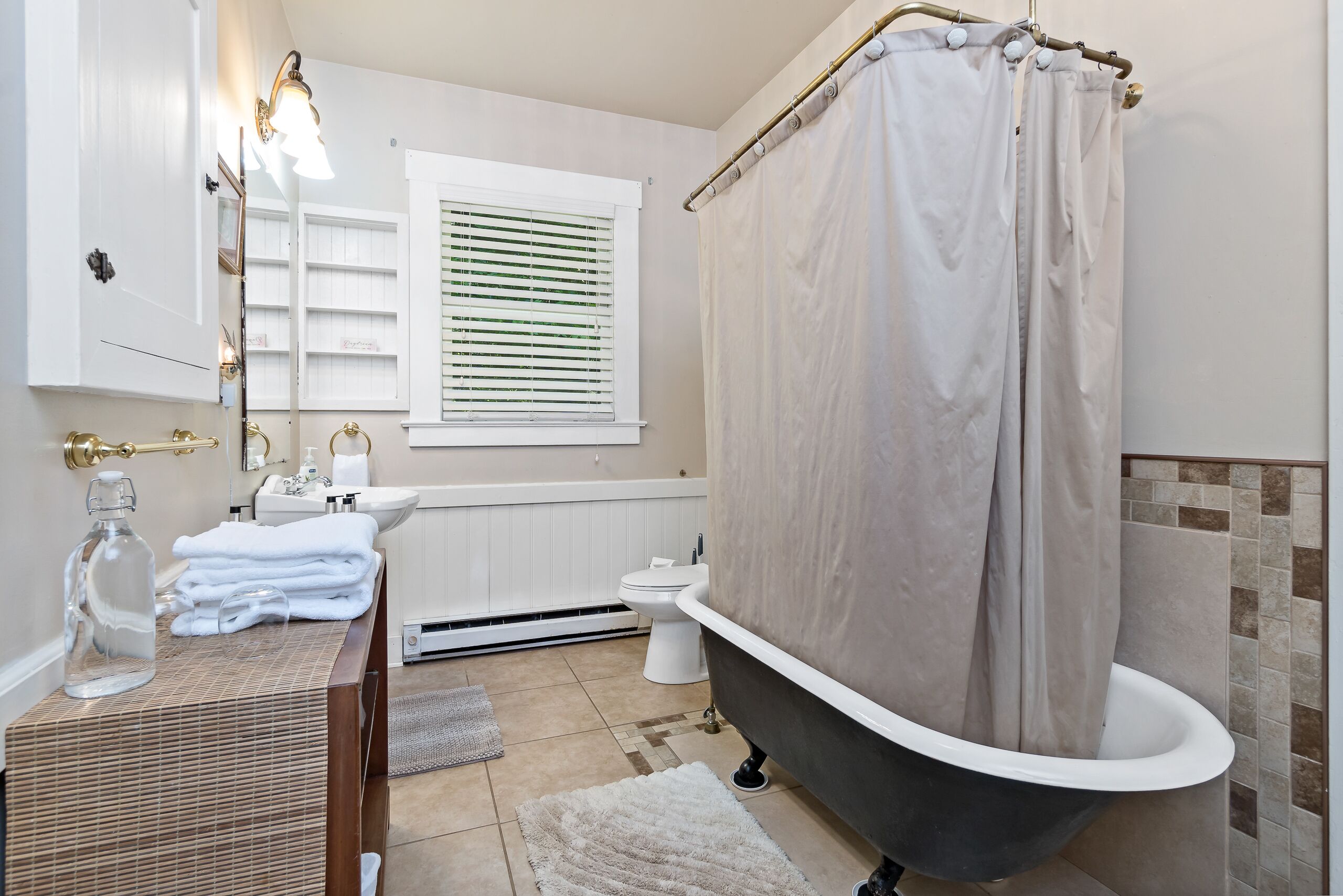Un baño con bañera, lavabo con espejo, ventana con persianas y toallero.