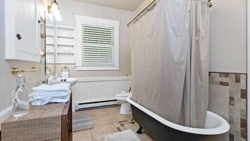 Un baño con bañera, lavabo con espejo, ventana con persianas y toallero.