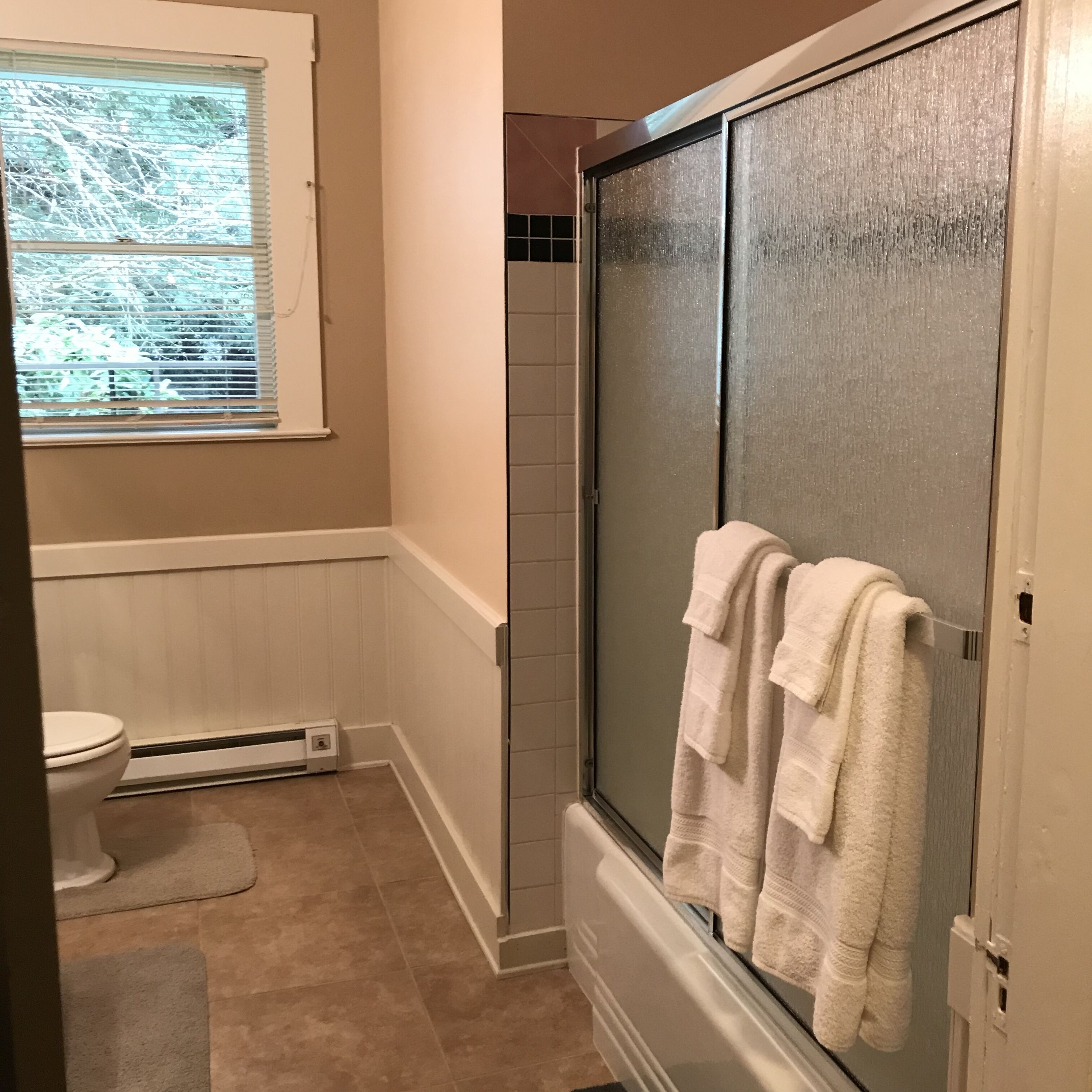 Ein Badezimmer mit Toilette, einem Fenster mit Jalousien, einer Dusche mit Glastür und einem Handtuch, das an der Tür hängt.
