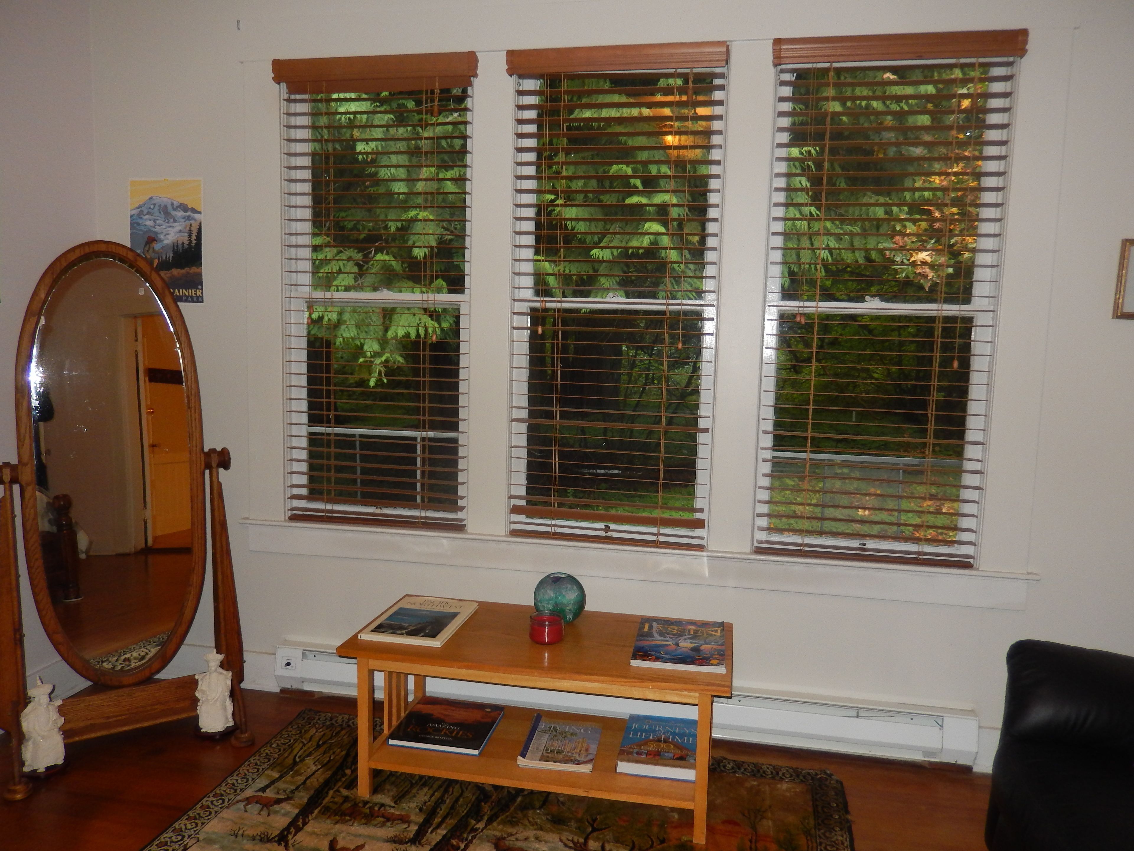 Una habitación con tres ventanas con persianas, una mesa de madera con libros, un espejo y una alfombra.