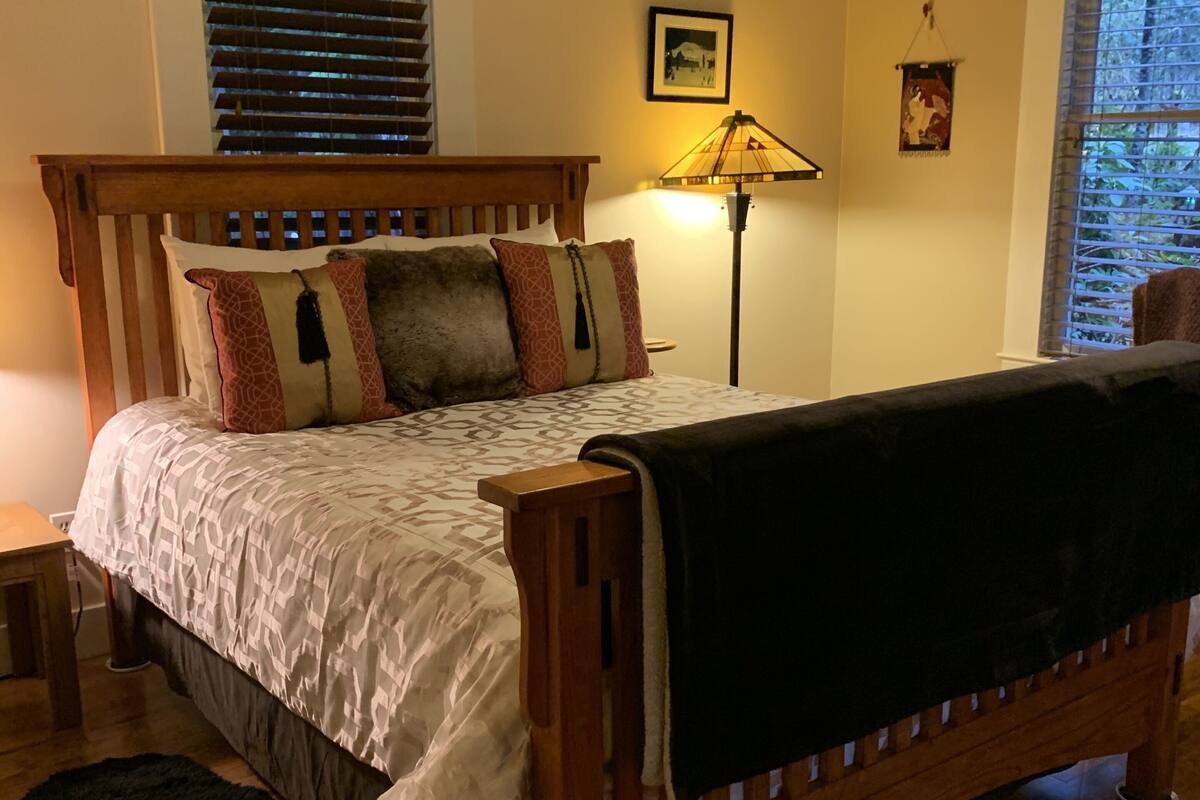 A wooden bed with a patterned bedspread and multiple pillows, a bedside lamp, a black rug, and a window with blinds.