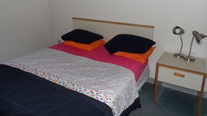 Un lit recouvert d’une courtepointe à pois colorés, avec deux oreillers noirs et un oreiller orange. Une table de chevet, sur laquelle se trouve une lampe et un tiroir, est placée à côté du lit. Le mur est décoré de deux tableaux jaunes.