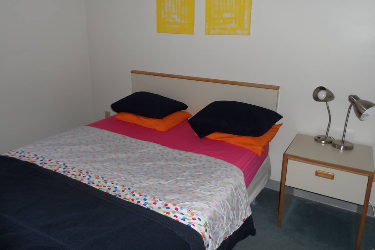 Ein Bett mit einem farbenfrohen, weißen Bettbezug mit Punkten, zwei schwarzen Kissen und einem orangefarbenen Kissen. Ein Nachttisch mit Lampe und Schublade steht daneben. An der Wand hängen zwei gelbe Bilder.