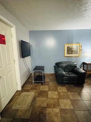 A room with a television mounted on the wall, a black leather sofa, a framed picture, and a tiled floor. - Economy Suites (Muldraugh)