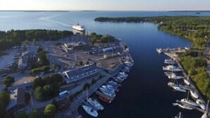 An aerial view of a marina with boats docked, a large ship in the distance, and a forested island.