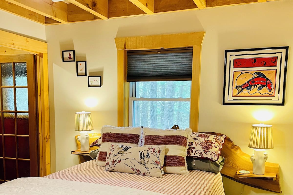 A bedroom with a large bed, wooden ceiling beams, a window with a view of trees, and a framed picture on the wall.
