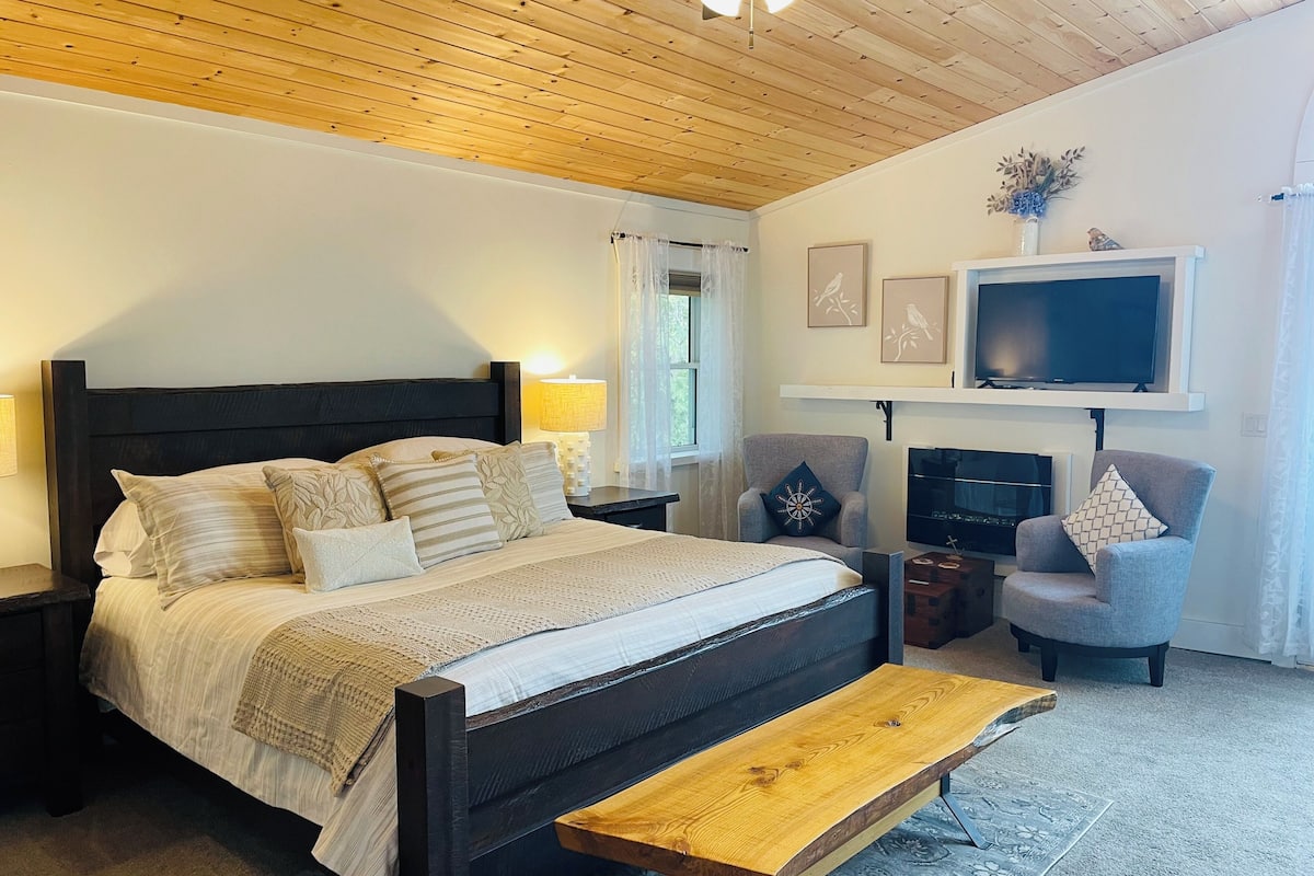 A bedroom with a bed, bedside tables, a TV, a fireplace, and a chair.