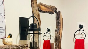 A bathroom with a wooden vanity, a stone sink, a mirror with a rustic frame, a red towel, and a small shelf with a candle and a framed quote.