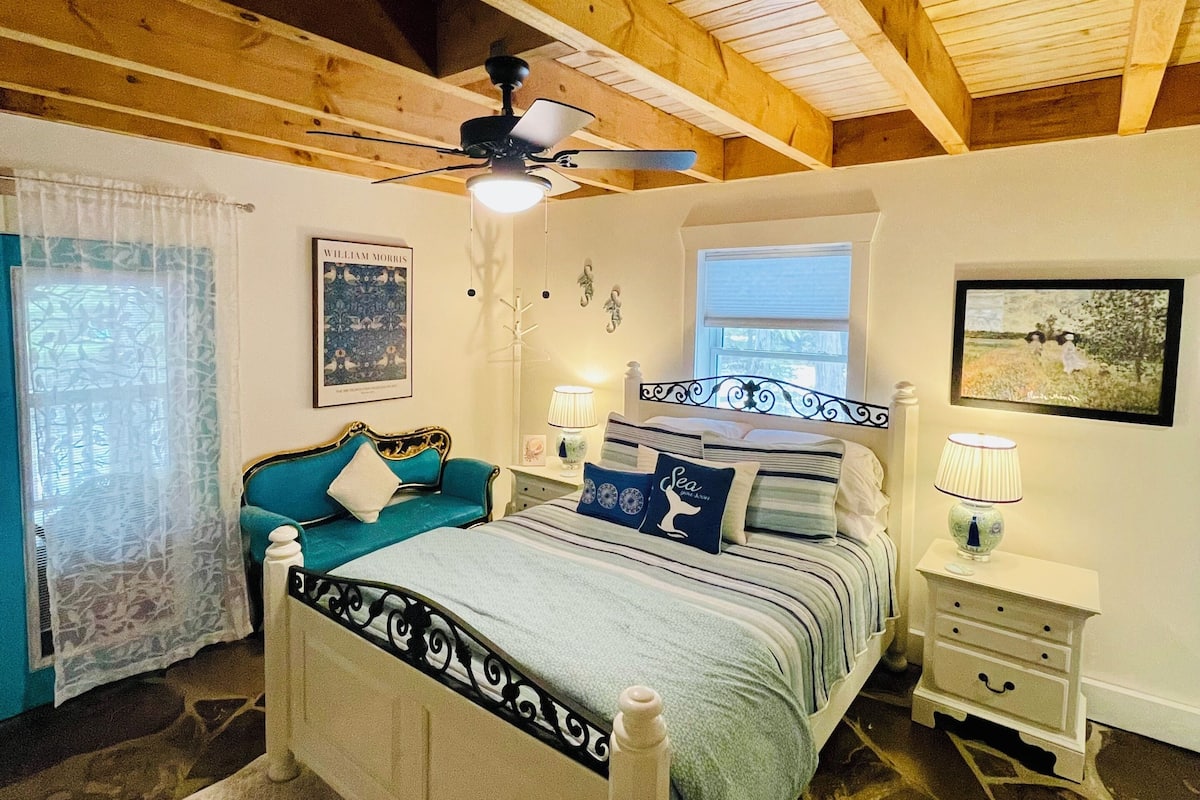 A bedroom with a wooden ceiling, a bed with a headboard, a nightstand, a lamp, and a window with curtains.