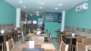 Un restaurant avec des tables et des chaises en bois, un espace cuisine à l’arrière-plan et un grand écran mural.