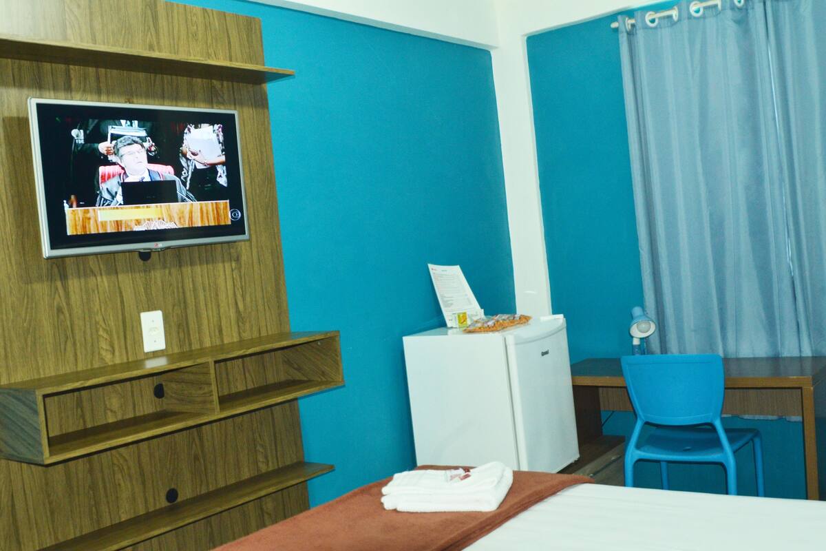 A hotel room with a television mounted on a wooden shelf, a small white refrigerator, a blue chair, and a bed with a brown headboard.