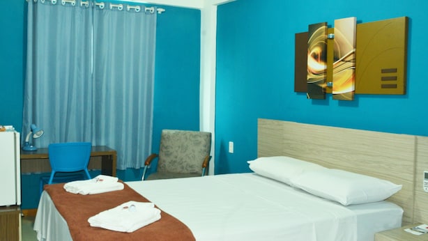 Une chambre d’hôtel avec un lit, un bureau et une chaise, un rideau bleu et des œuvres d’art accrochées au mur.