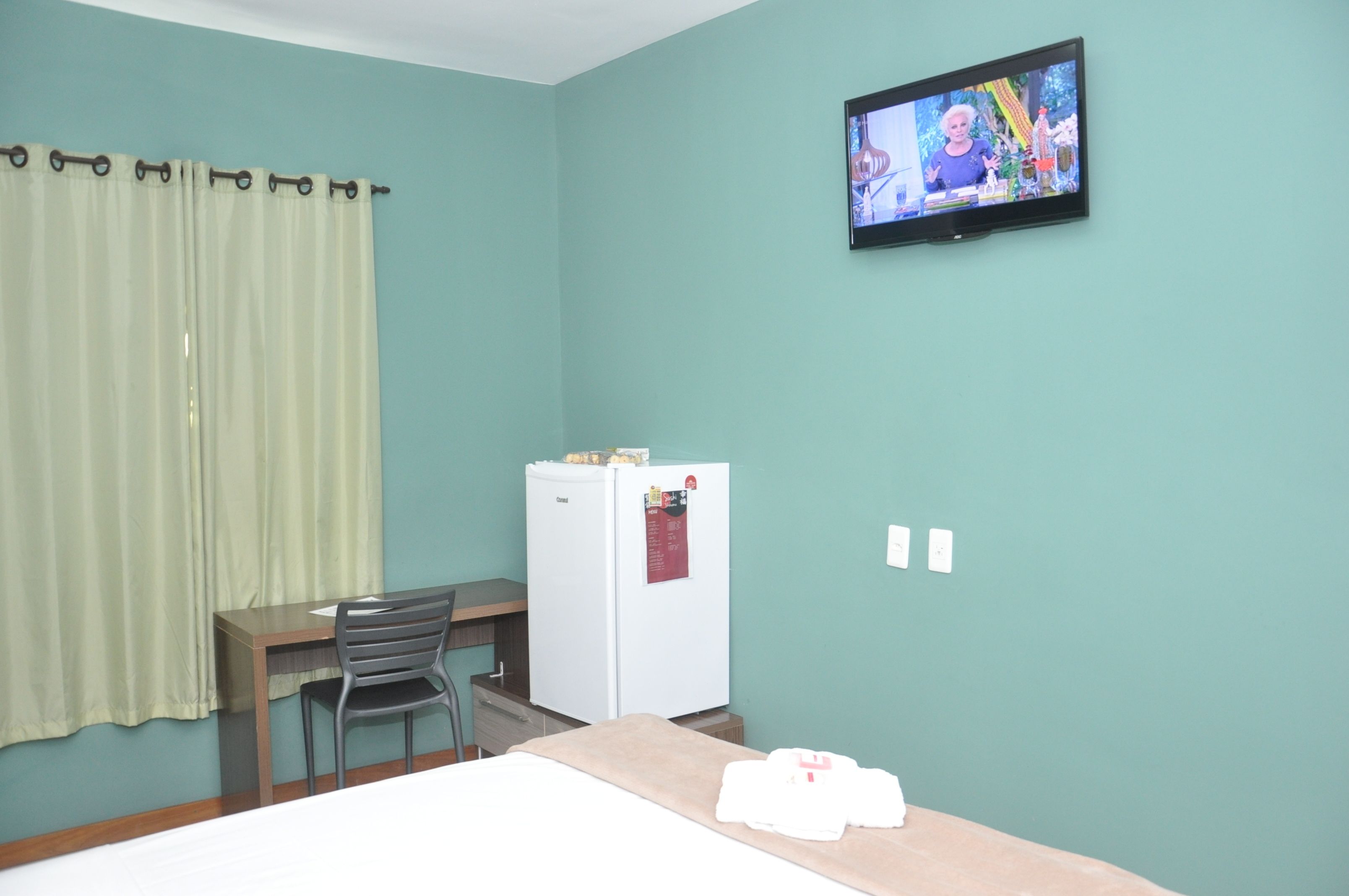 Una habitación con paredes color esmeralda, una cama, un escritorio con una silla, un refrigerador blanco y un televisor en la pared.