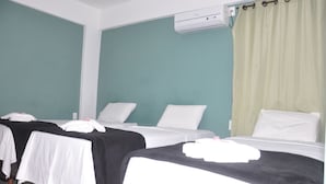 Une chambre d’hôtel avec trois lits, chacun étant équipé de draps blancs et d’une couverture noire. On y trouve un climatiseur au plafond et un rideau vert à la fenêtre.