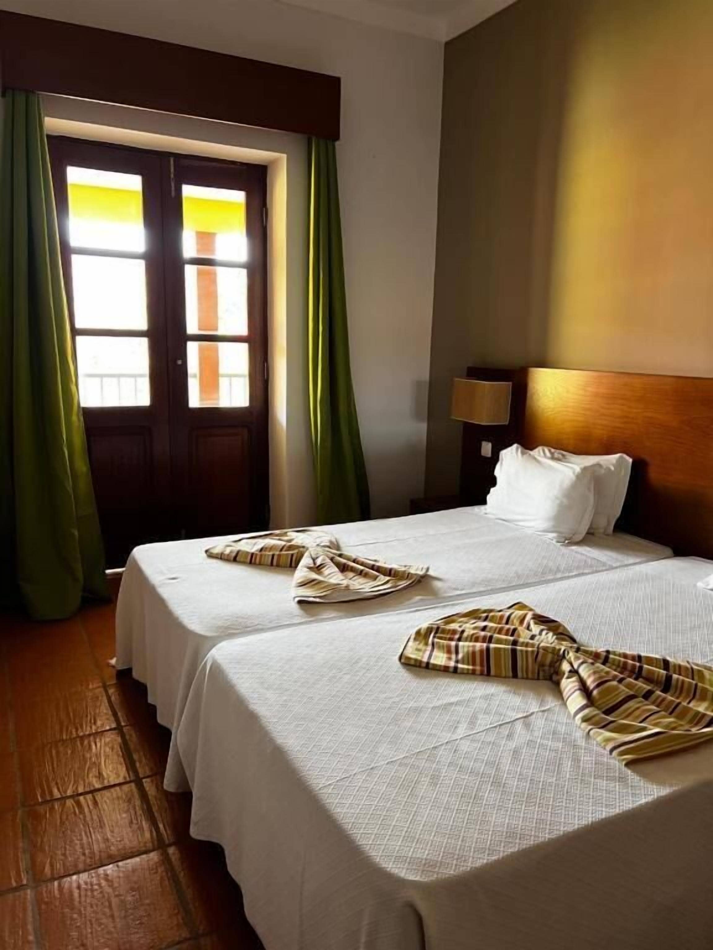 A hotel room with two beds, a wooden headboard, a nightstand, and a window with curtains.