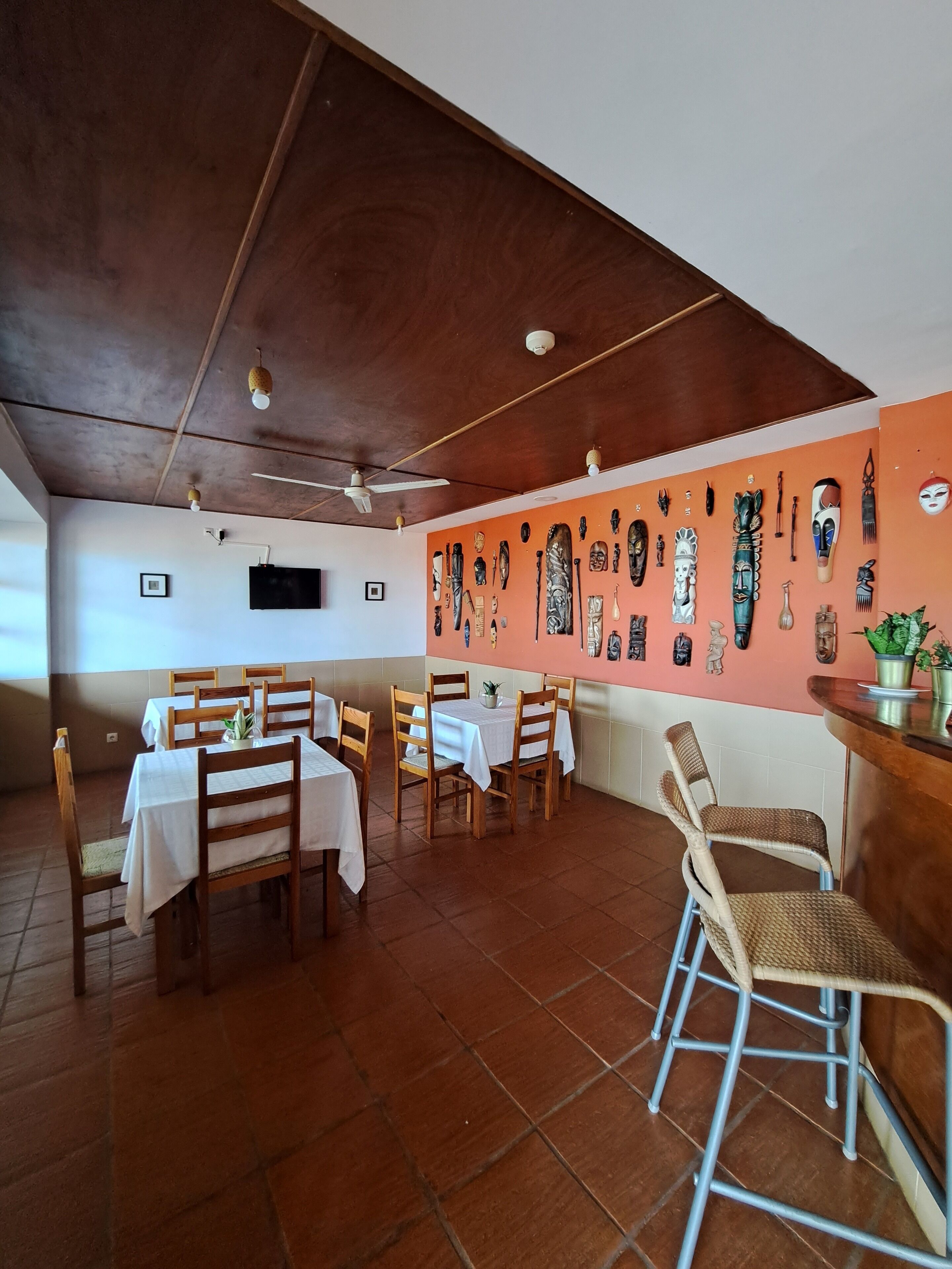A restaurant with wooden tables and chairs, a wall of masks, and a ceiling fan.