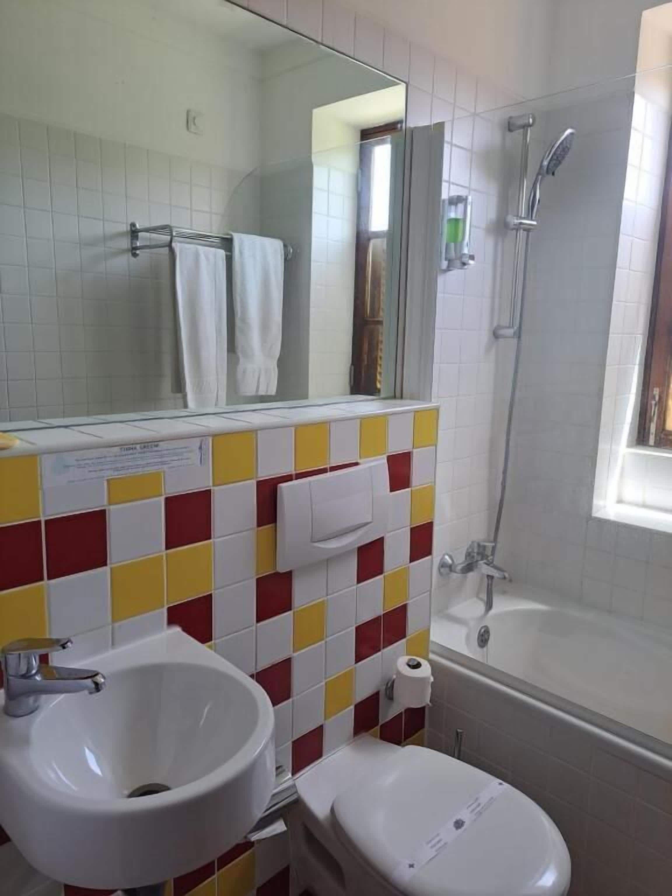A bathroom with a white sink, toilet, and shower. The walls are tiled in a red and yellow pattern.