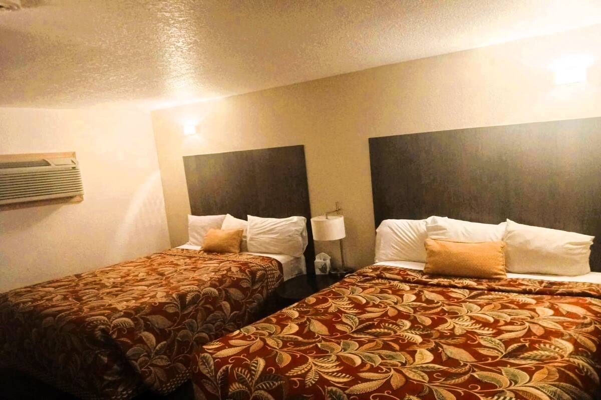 A hotel room with two beds, each with a patterned bedspread, a bedside lamp, and a wall-mounted air conditioner.