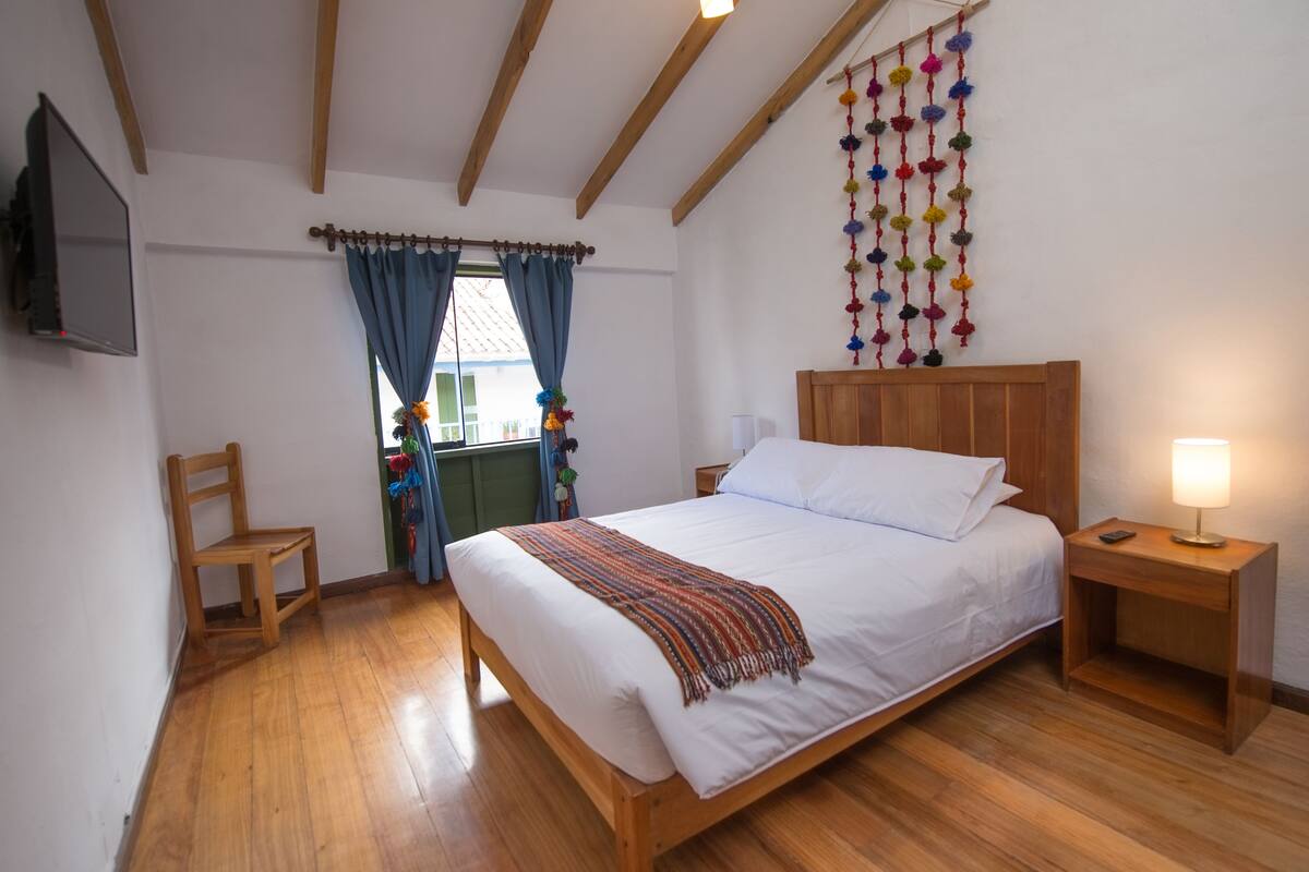 A bedroom with a wooden bed, a nightstand with a lamp, a chair, and a TV mounted on the wall.