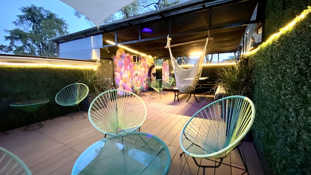 En udendørs patio med runde glastop borde og grønne wirestole, et overdækket loungeområde med hængestole, samt en væg med et farverigt mural.