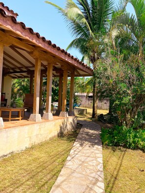 A covered outdoor area with a stone pathway, wooden furniture, and lush greenery including palm trees.