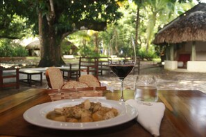 Una mesa de comedor con un plato de comida, una copa de vino tinto y un vaso de agua, ubicada en un espacio al aire libre con árboles y una estructura de techo de paja.