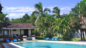 Una piscina con sillas azules, un edificio pequeño con techo de tejas y palmeras.
