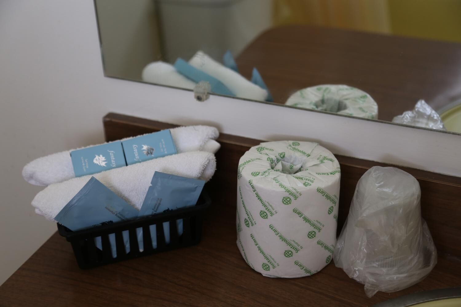 A hotel room with a wooden headboard, a black basket containing folded towels, a toilet paper roll, and a plastic bag.