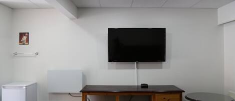 A room with a flat-screen TV mounted on the wall, a wooden desk with drawers, a small round table, and a white radiator.