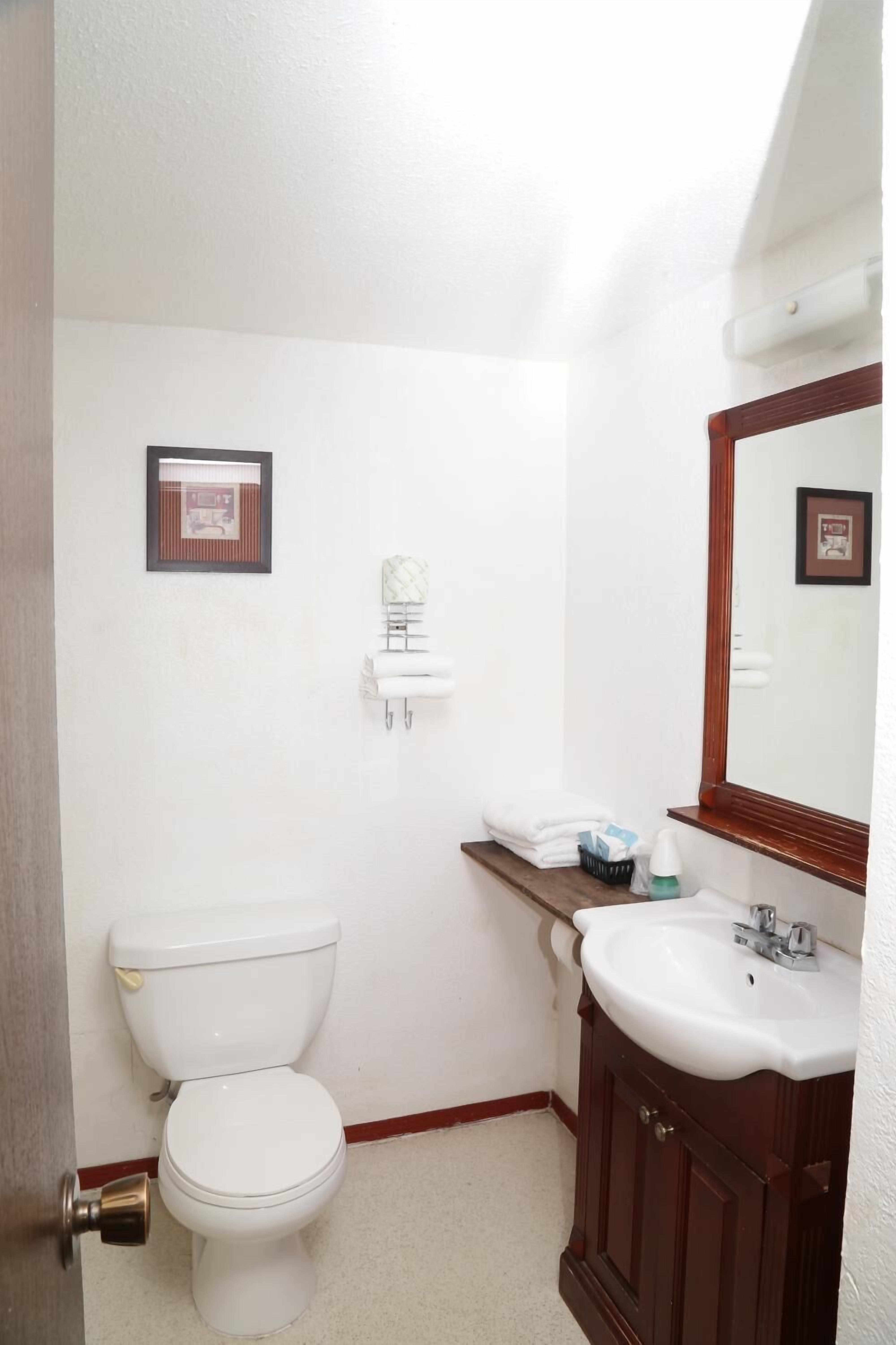 Une petite salle de bain propre, avec des toilettes, un lavabo et un miroir. Les murs sont ornés de tableaux encadrés et il y a un porte-serviettes.