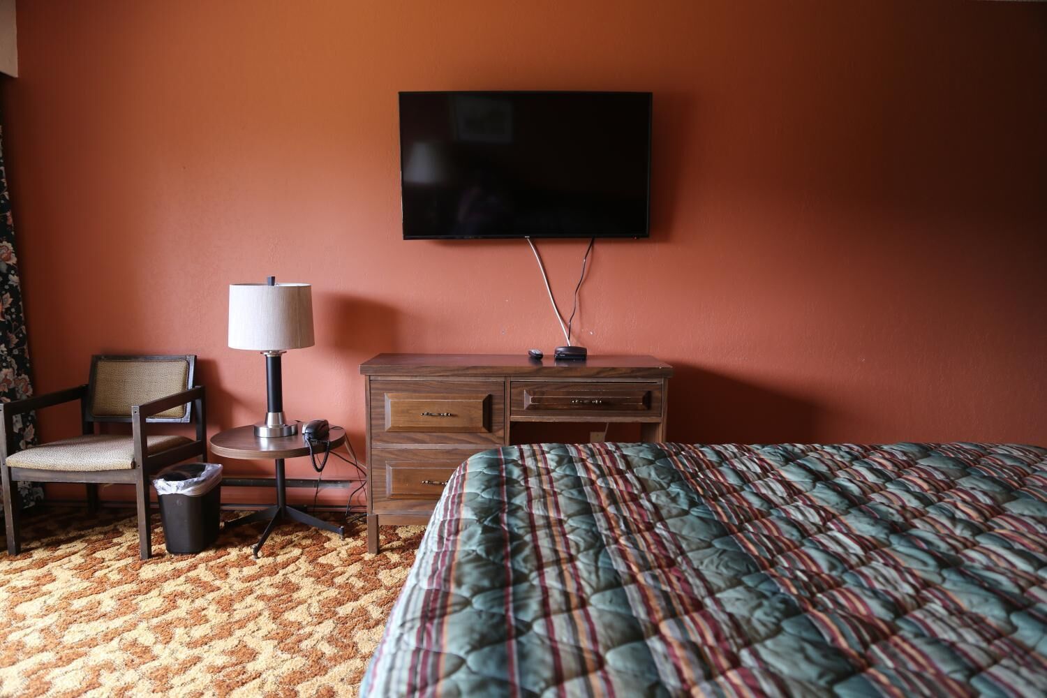 A hotel room with a bed, a wooden nightstand, a chair, a lamp, and a television mounted on the wall.