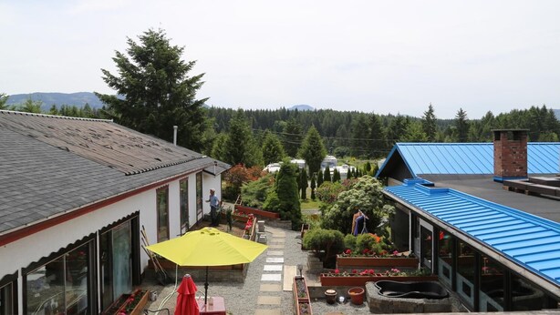 A courtyard with a building featuring a sloped roof, a yellow umbrella, and a red table. - Timberlodge RV & Campground (Port Alberni)