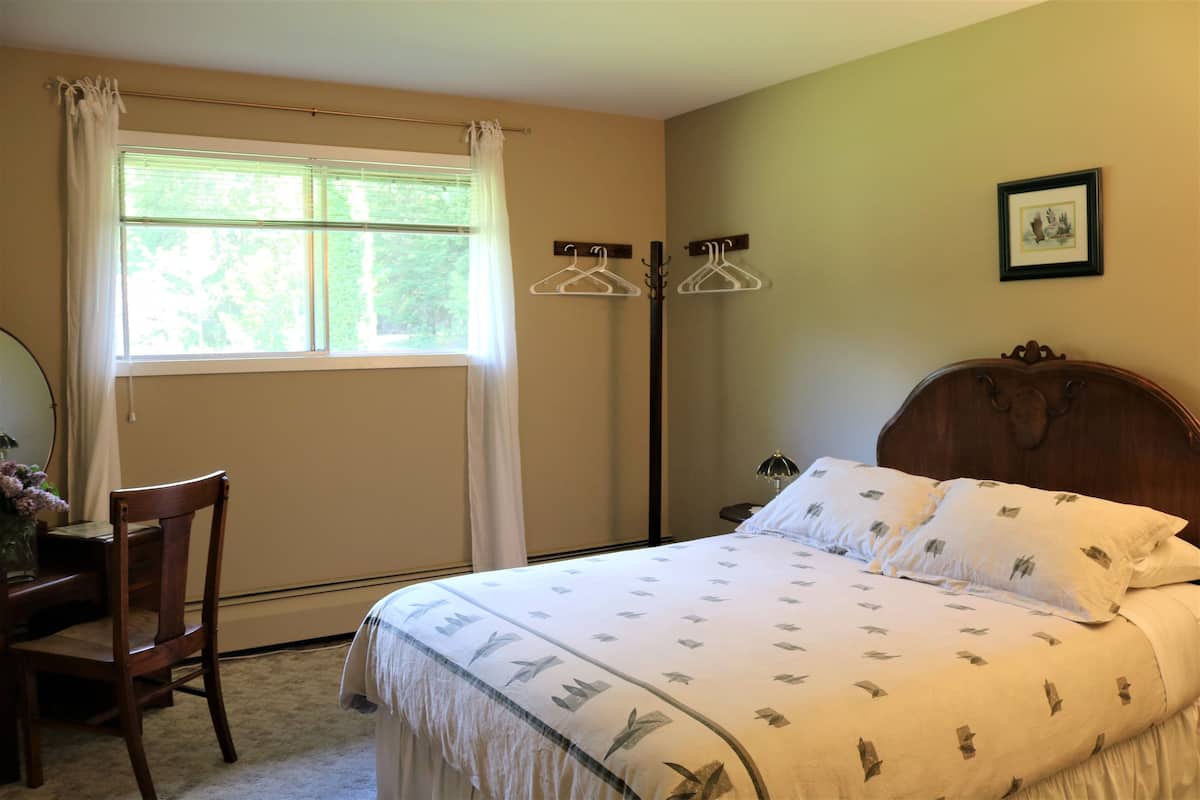 Quarto com cama, cadeira, escrivaninha, janela com cortinas e um cabideiro.