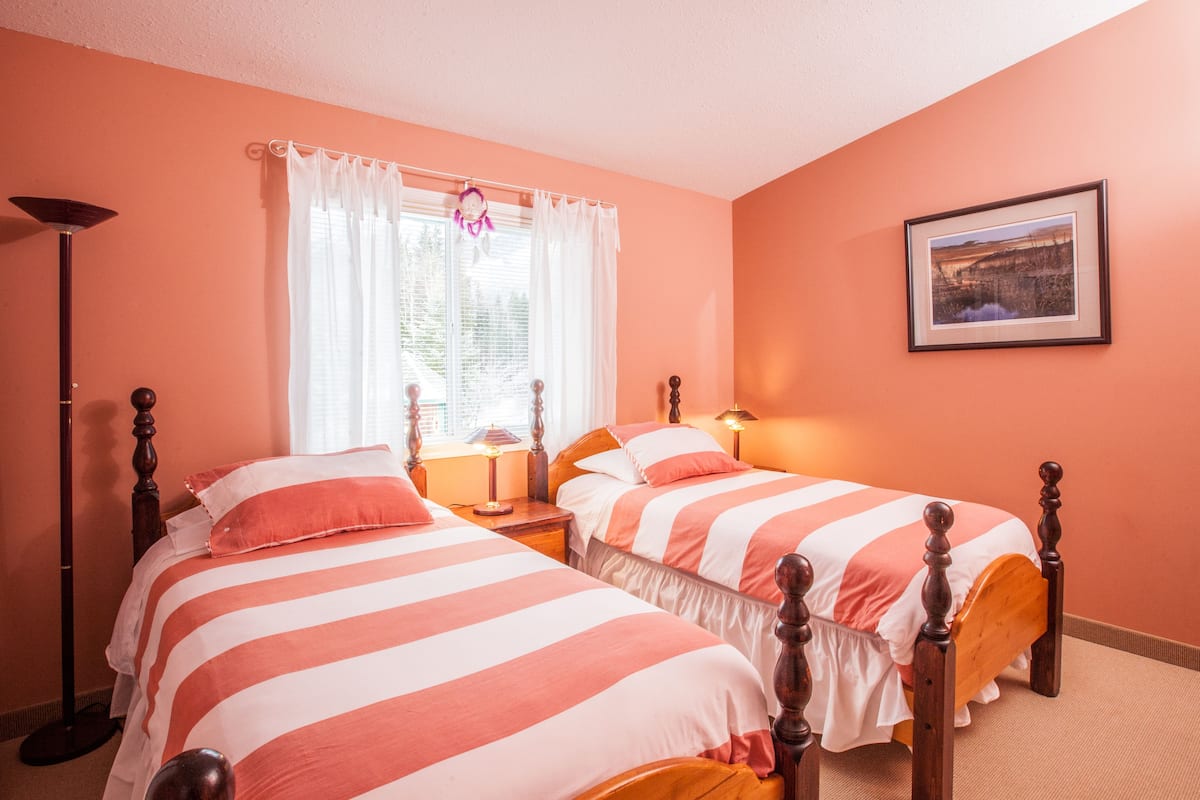 Quarto com duas camas, janela com cortinas, abajur e quadro na parede.