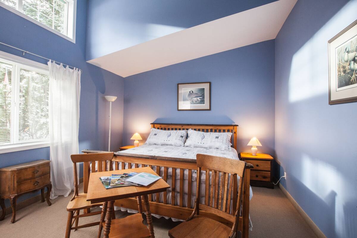 Quarto com cama, mesas de cabeceira, uma cadeira de madeira, uma mesa com uma revista e uma janela com cortinas.