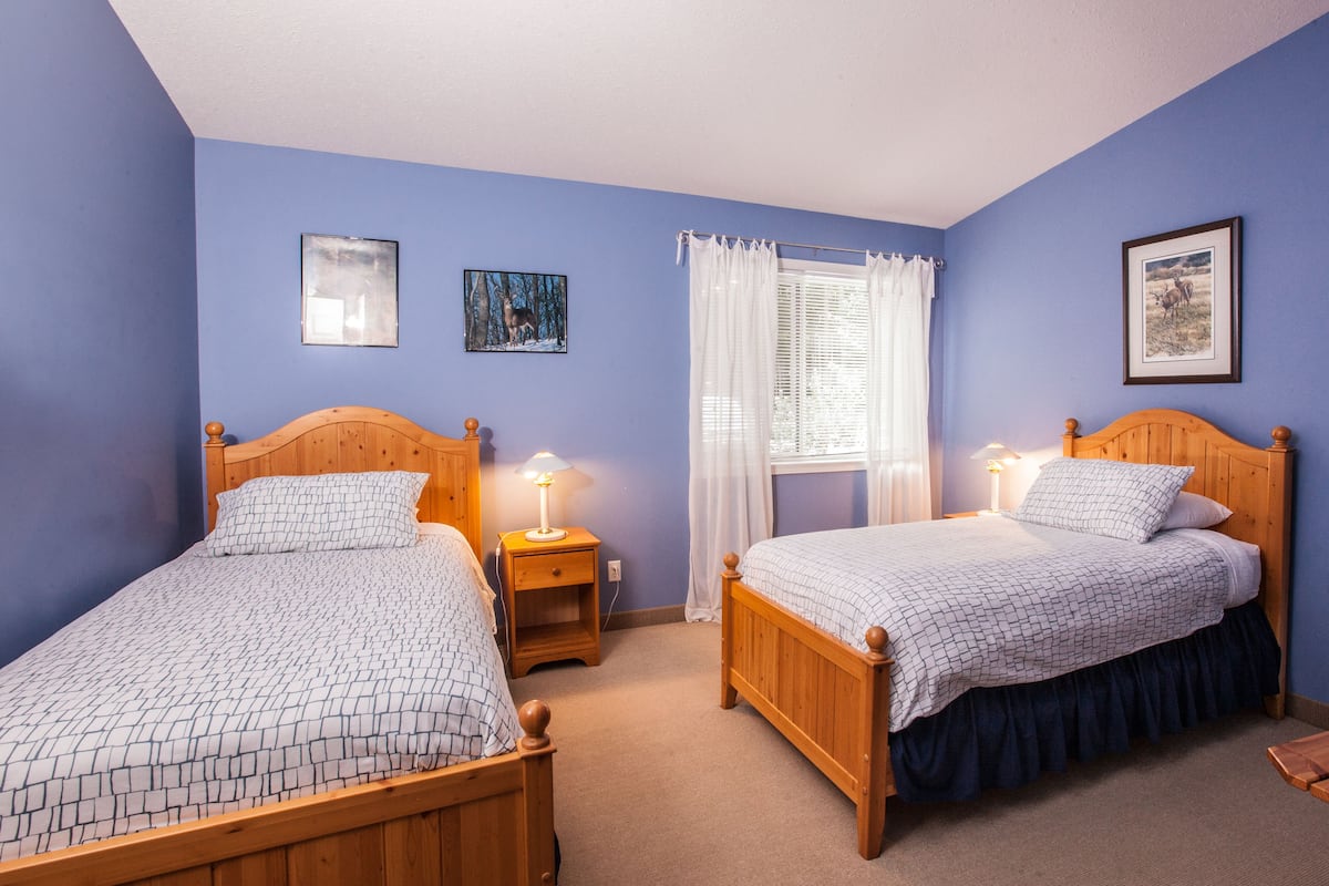 Quarto com duas camas de madeira, uma janela com cortinas brancas e uma pequena mesa de madeira.