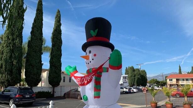 Una decoración de muñeco de nieve en un estacionamiento, con cerca de piquetes blancos, coches aparcados y edificios al fondo.