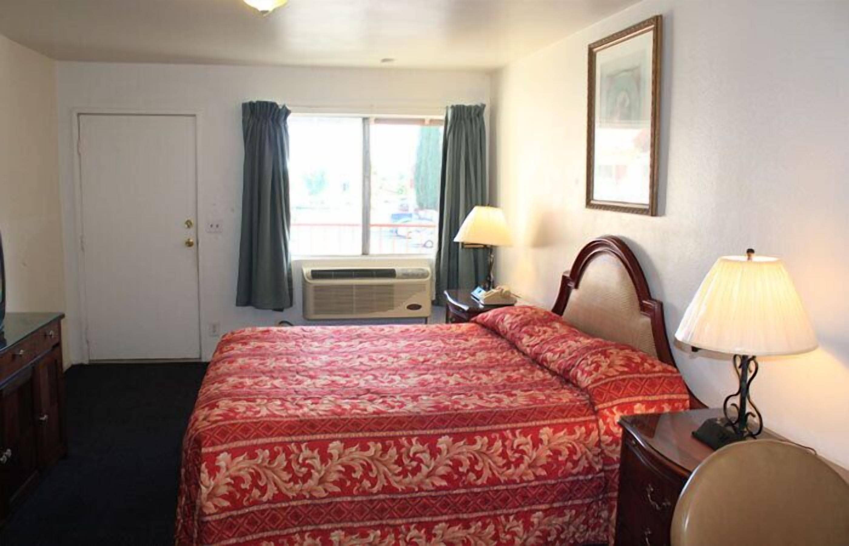 Habitación de hotel con cama, escritorio, silla, lámpara, ventana con cortinas, unidad de aire acondicionado y puerta.