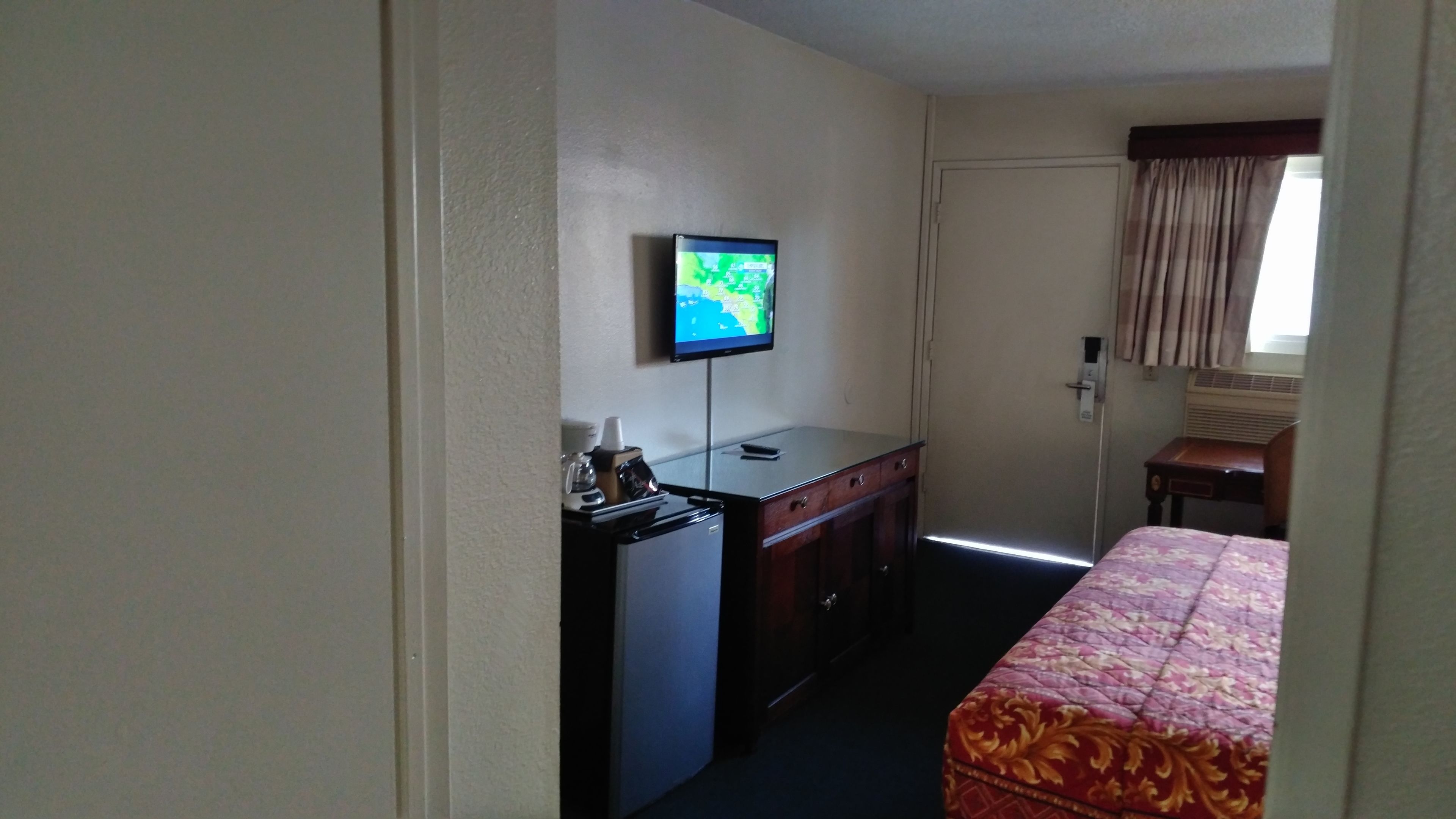 Una habitación de hotel con cama, escritorio con computadora, televisor montado en la pared y ventana con cortinas.