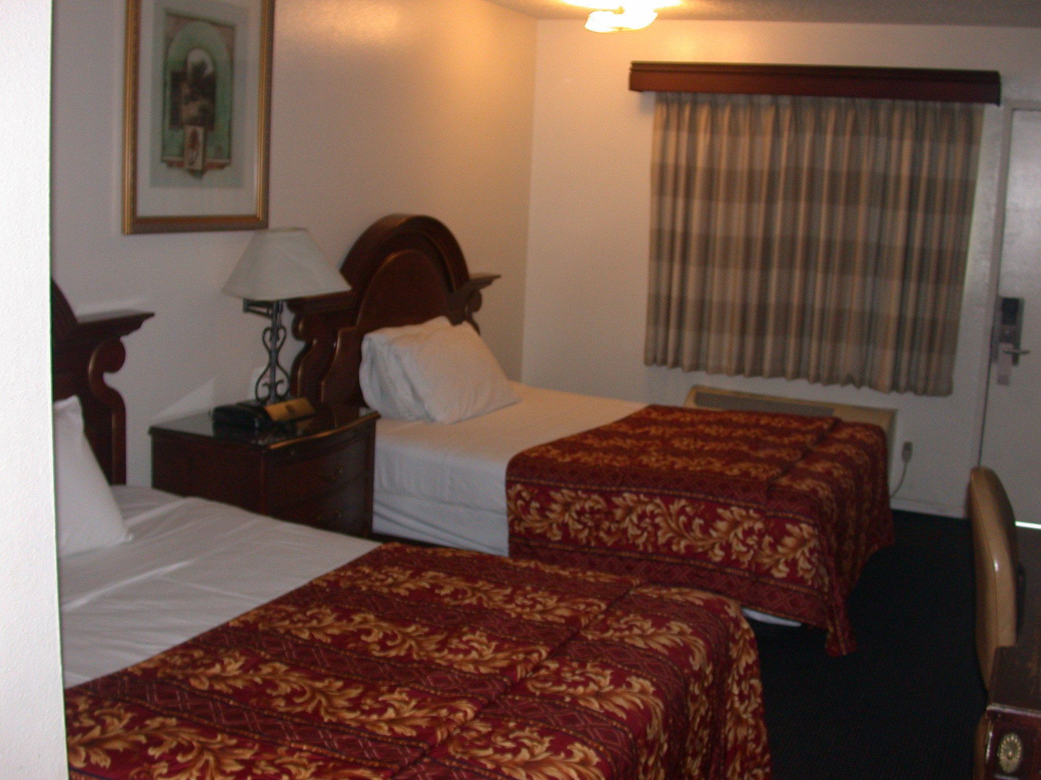 A hotel room with two single beds, a nightstand with a lamp, a wooden headboard, a window with curtains, and a chair.
