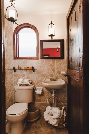 A bathroom with a stone tile wall, a wooden-framed window, a white sink, a toilet, and a basket of towels.